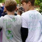 Zwei Jungen tragen Partner-T-Shirts. Auf dem Linken steht "Helft uns", auf dem Rechten steht "Das Klima zu retten."