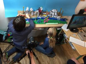 Vor einem Tisch sitzen zwei Kinder und bauen eine Fantasielandschaft mit Bergen und Lebewesen aus Pfeifenanzündern auf. Eine Kamera steht bereit, um die aufgebaute Szene zu fotografieren.